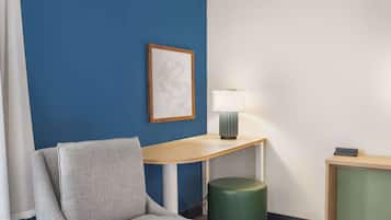 Una habitación con una silla tapizada de gris, una mesita de madera con una lámpara, una obra enmarcada en la pared azul y un objeto cilíndrico verde.