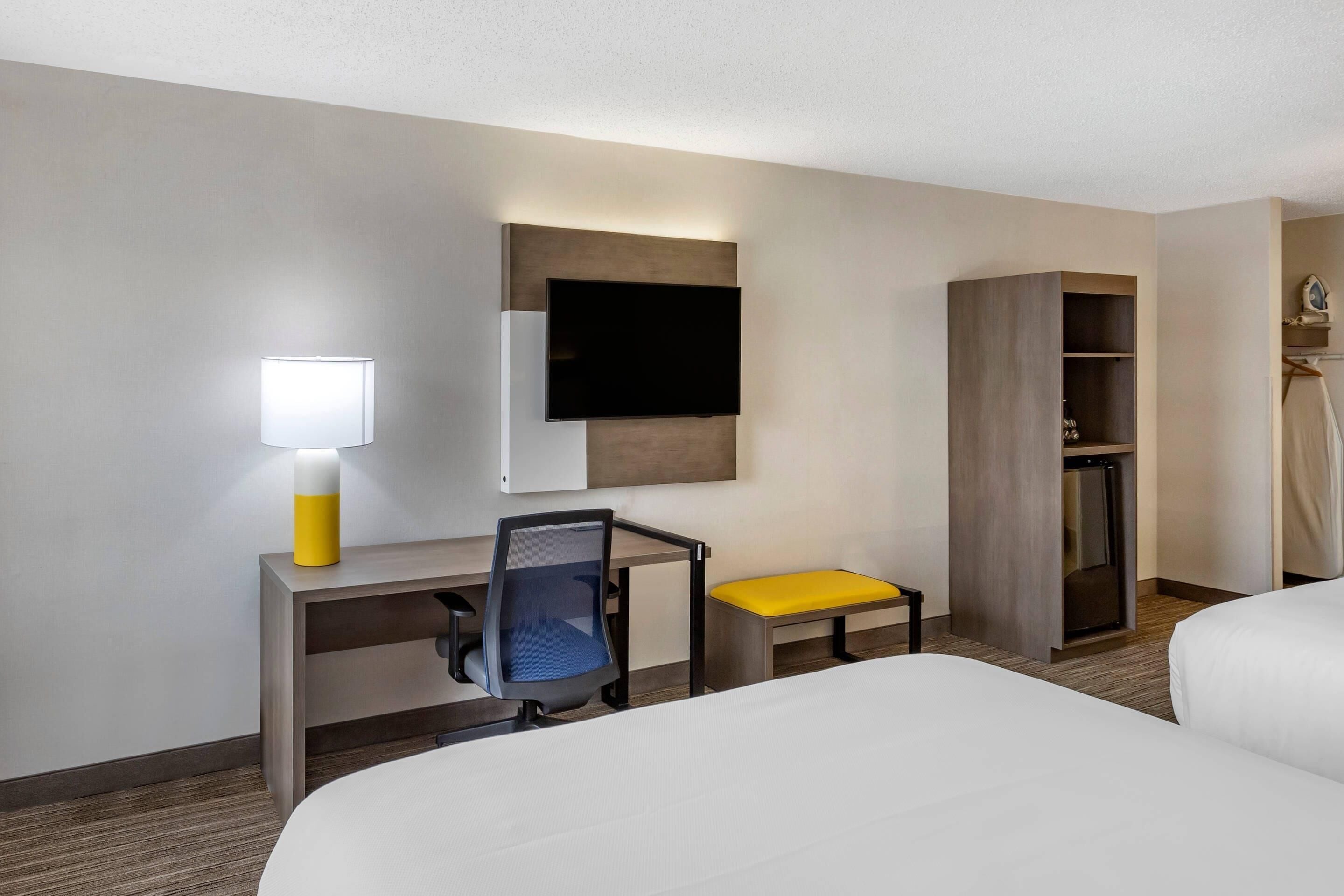 Habitación de hotel con cama, escritorio con silla, televisor montado en la pared, mesita de noche con lámpara y armario.