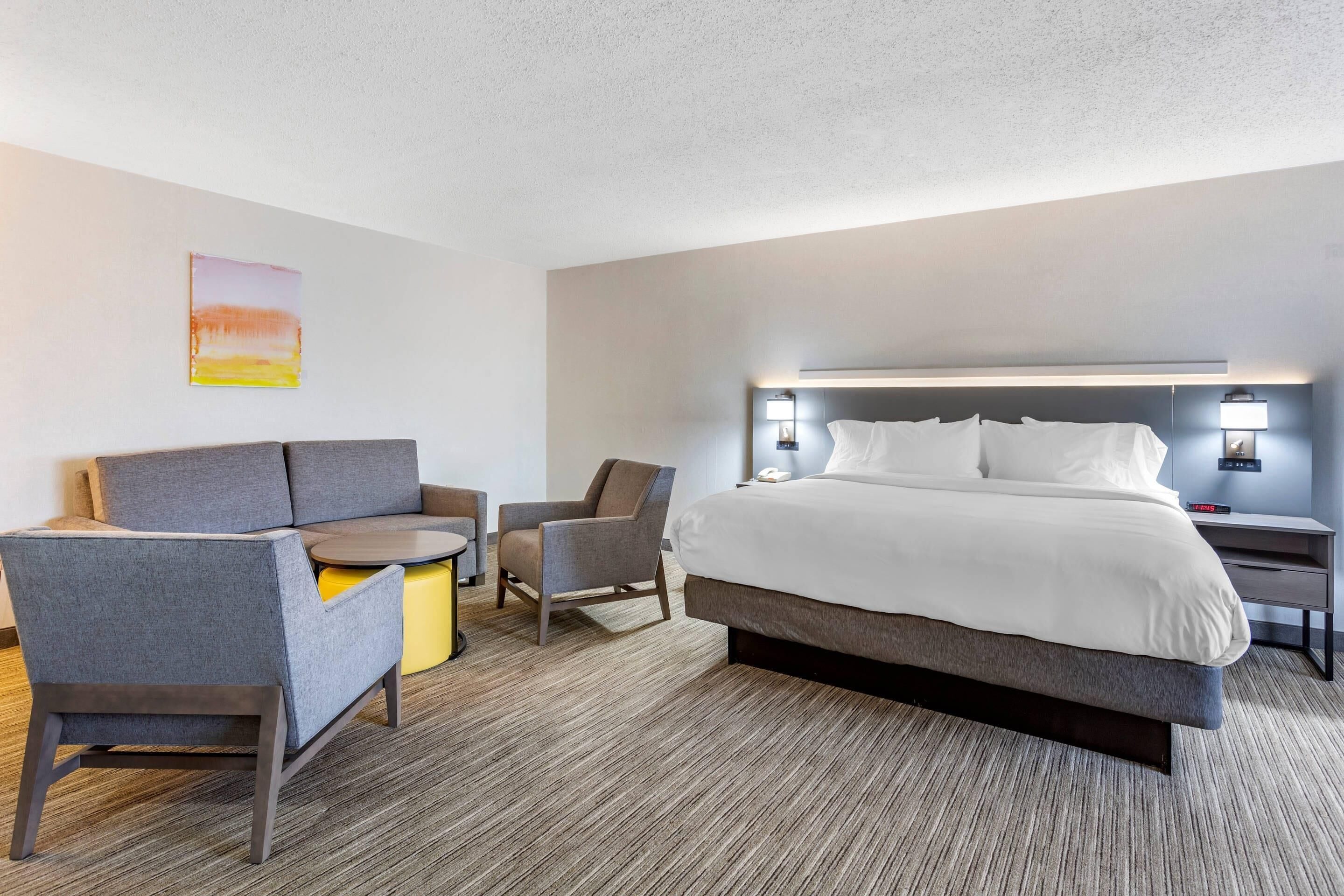 A hotel room with a bed, two armchairs, a small table, a nightstand, and a wall-mounted lamp.