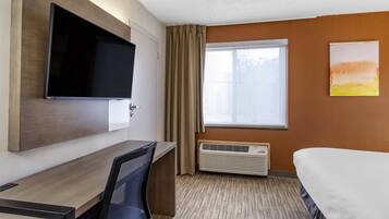 Habitación de hotel con cama, escritorio con silla, televisor montado en la pared, ventana con cortinas y radiador.