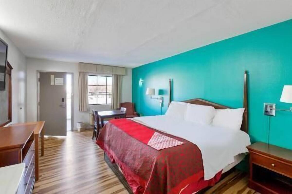 A hotel room with a bed, a desk, a chair, a lamp, and a window.
