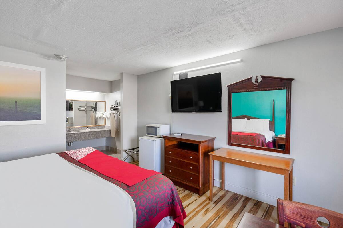 A hotel room with a bed, a desk, a TV, a mirror, and a small kitchenette.