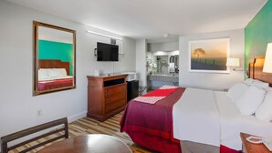 Una habitación de hotel con cama, escritorio, silla, espejo, televisor, un cuadro y baño a la vista.