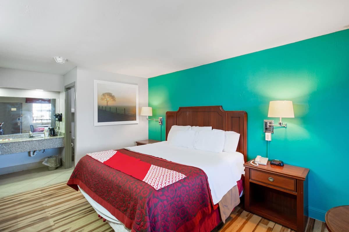 A hotel room with a bed, a nightstand, a lamp, a mirror, and a bathroom visible in the background.