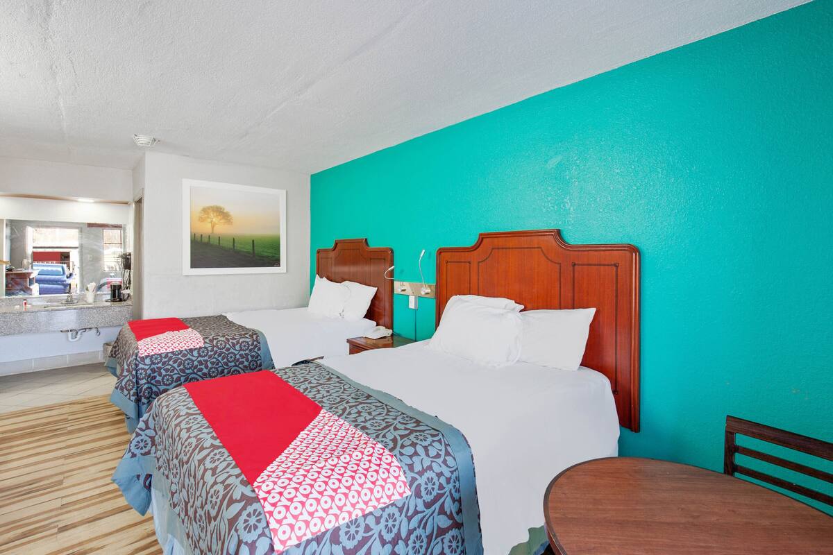 A hotel room with two beds, a wooden headboard, a teal accent wall, a painting, and a small table.