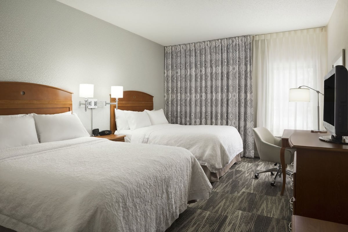 A hotel room with two beds, a desk, a chair, a TV, and a lamp.