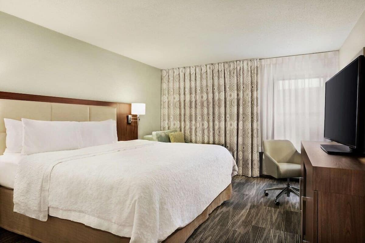 A hotel room with a large bed, a desk with a television, a chair, and a window with curtains.