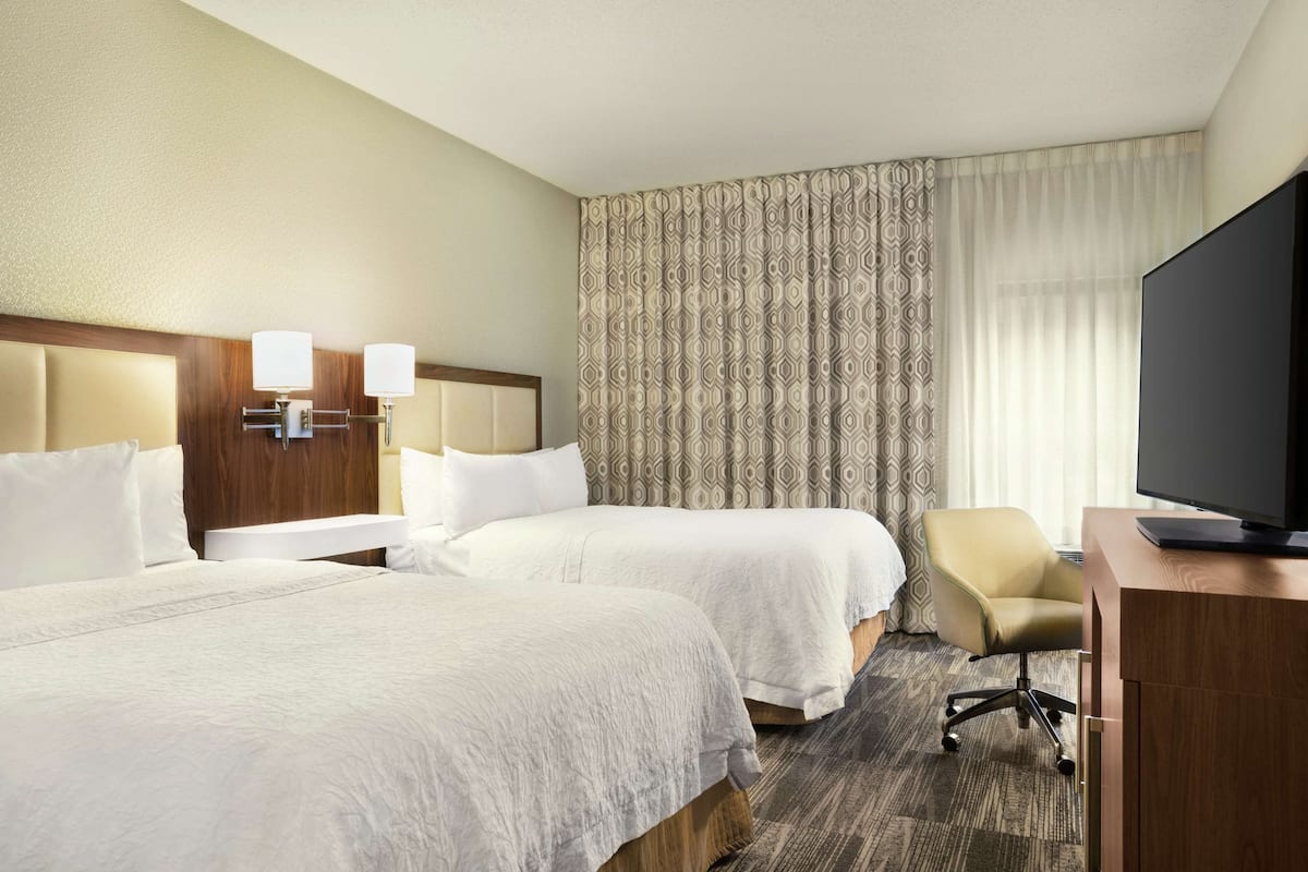 A hotel room with two beds, a desk with a flat-screen TV, a chair, and a patterned curtain.