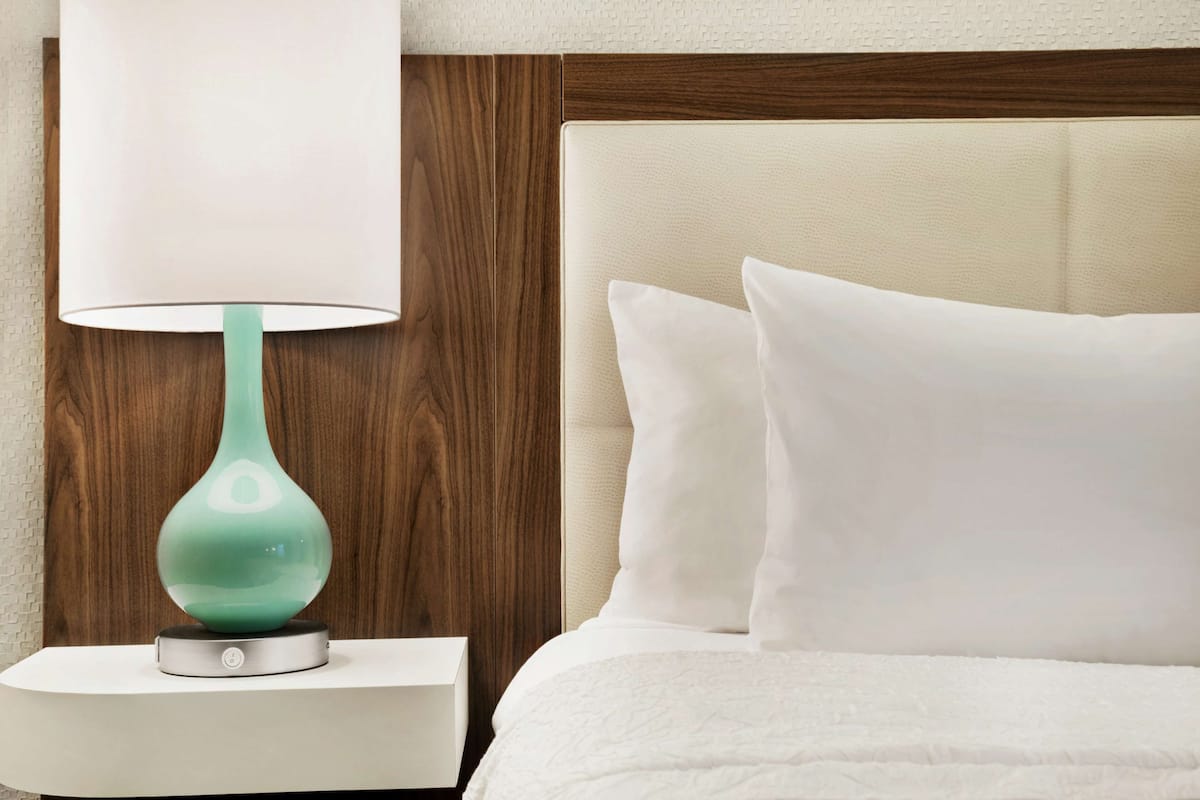 A neatly made bed with white bedding and pillows, a teal-colored table lamp on a white bedside table, and a wooden headboard.