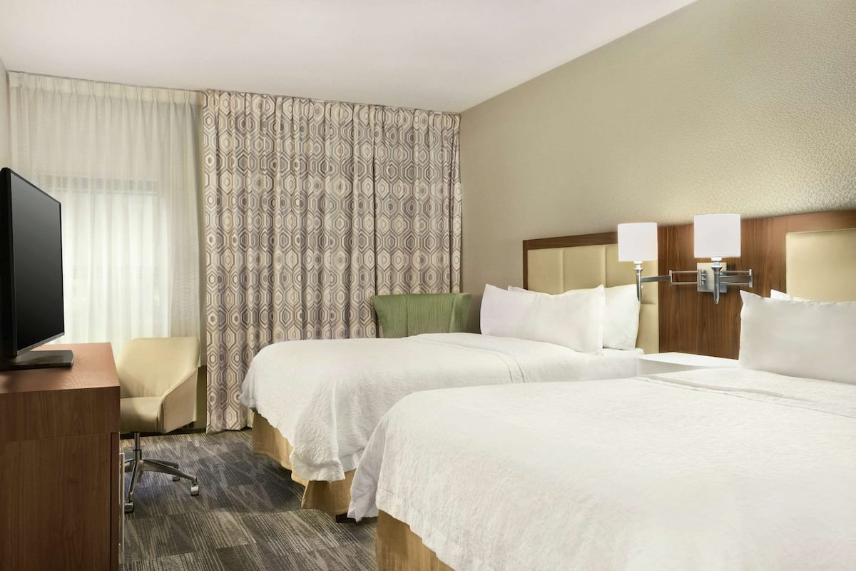 A hotel room with two beds, a desk with a television, a chair, and a patterned curtain.