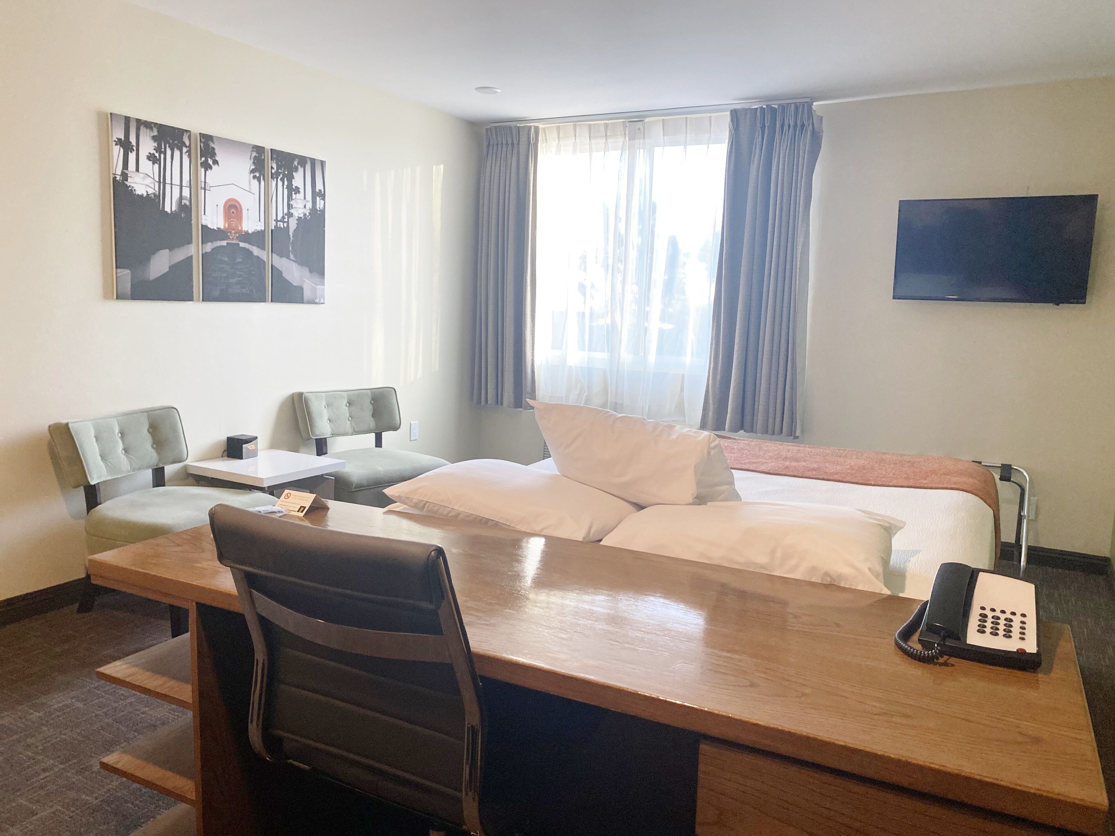 A hotel room with a bed, a desk, two chairs, a telephone, and a painting on the wall.