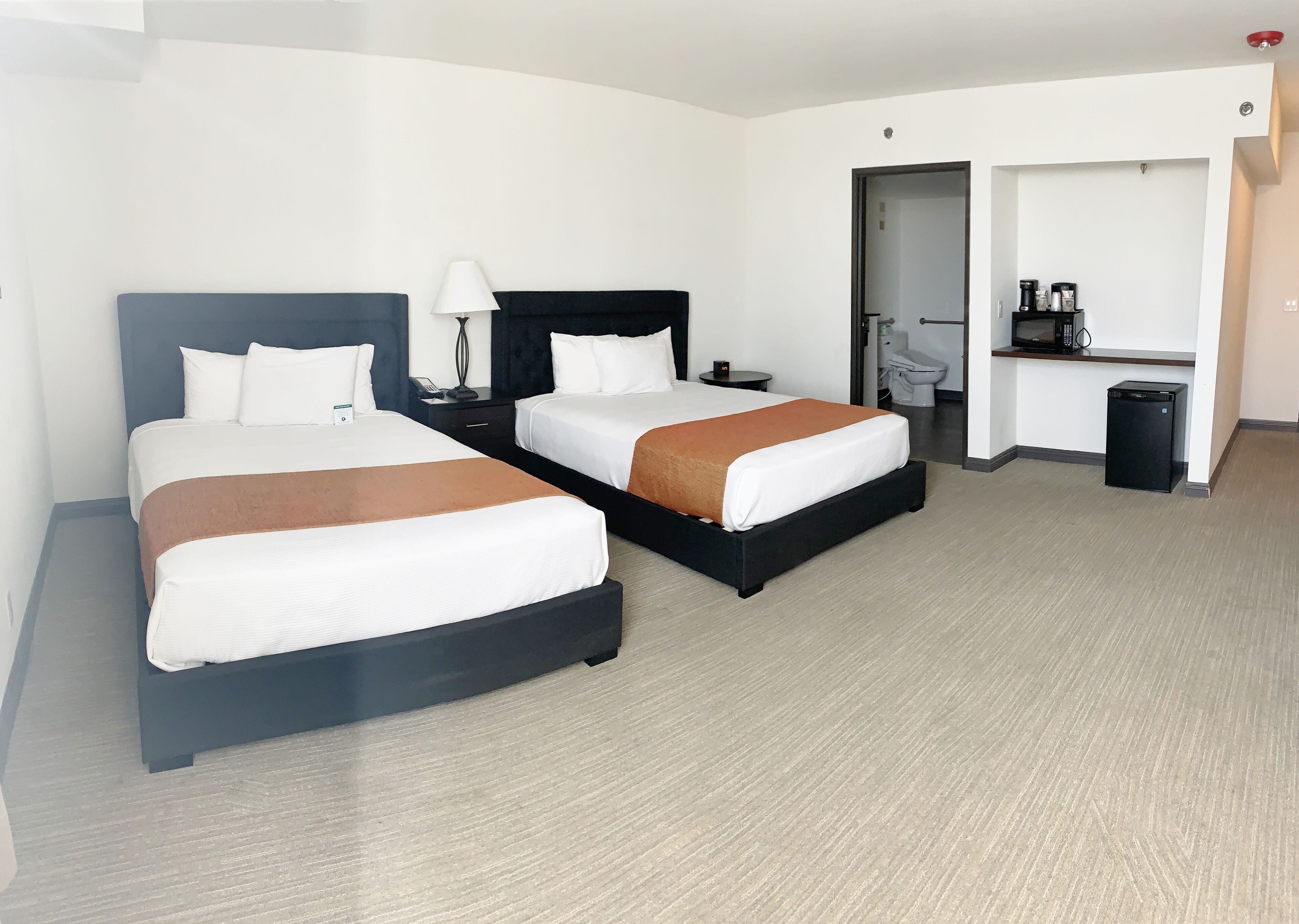 A hotel room with two beds, a desk with a computer, a trash bin, and a bathroom visible through an open door.