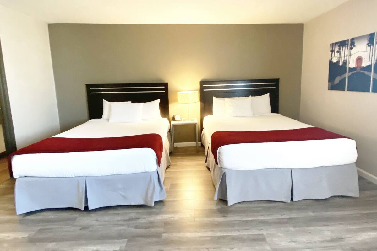 A hotel room with two beds, each with white bedding and red accents, a small bedside table with a lamp, and a wooden floor.