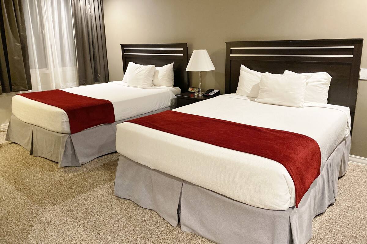 A hotel room with two beds, each with a red and white bedspread, a bedside table with a lamp, and a window with curtains.
