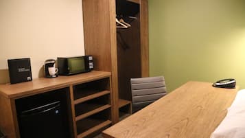 A hotel room with a wooden desk, a microwave, a coffee maker, a black suitcase, and a wooden wardrobe with a hanging rack.