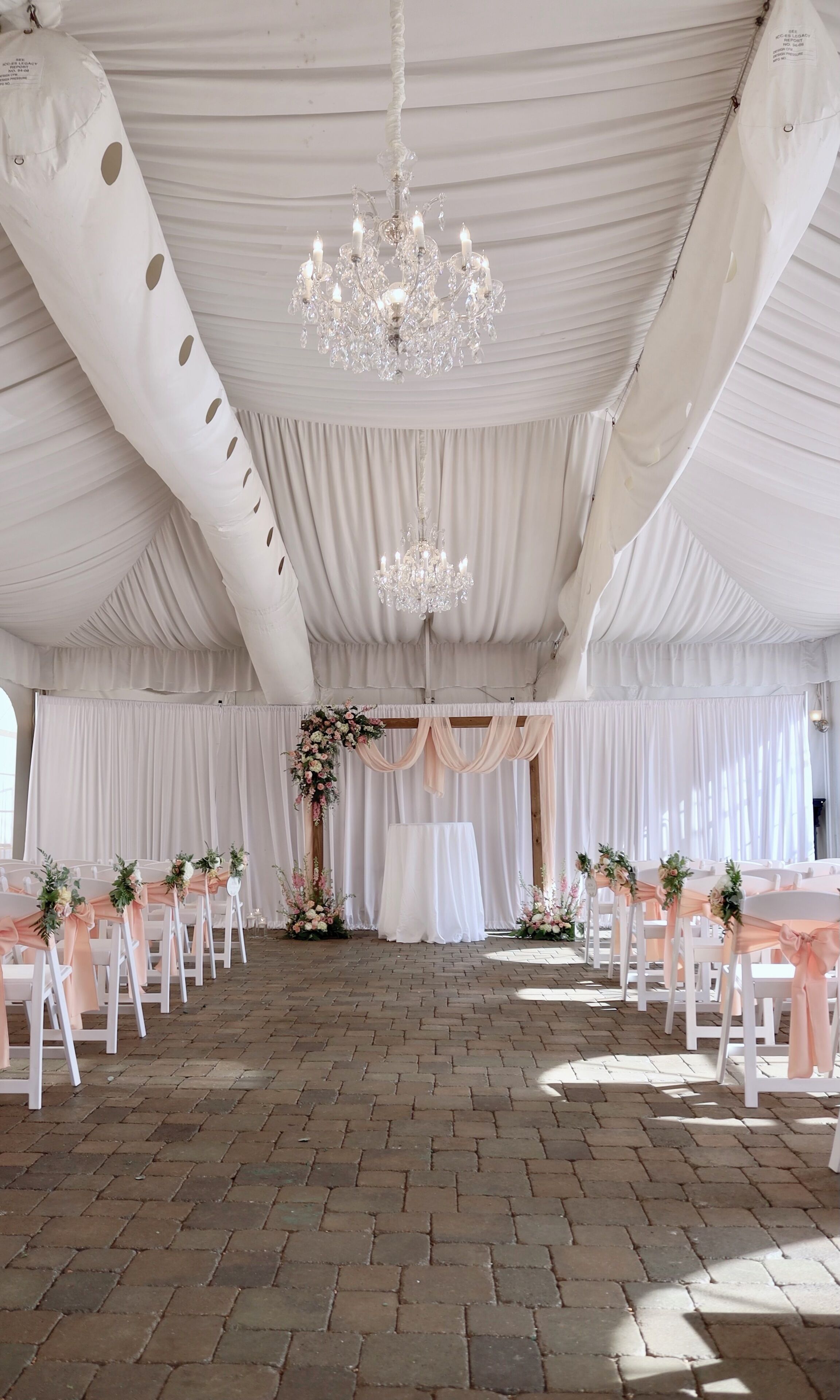 Un espace de réception pour mariage aménagé sous une tente, avec des tentures blanches, un lustre et des compositions florales en guise de centre de table.