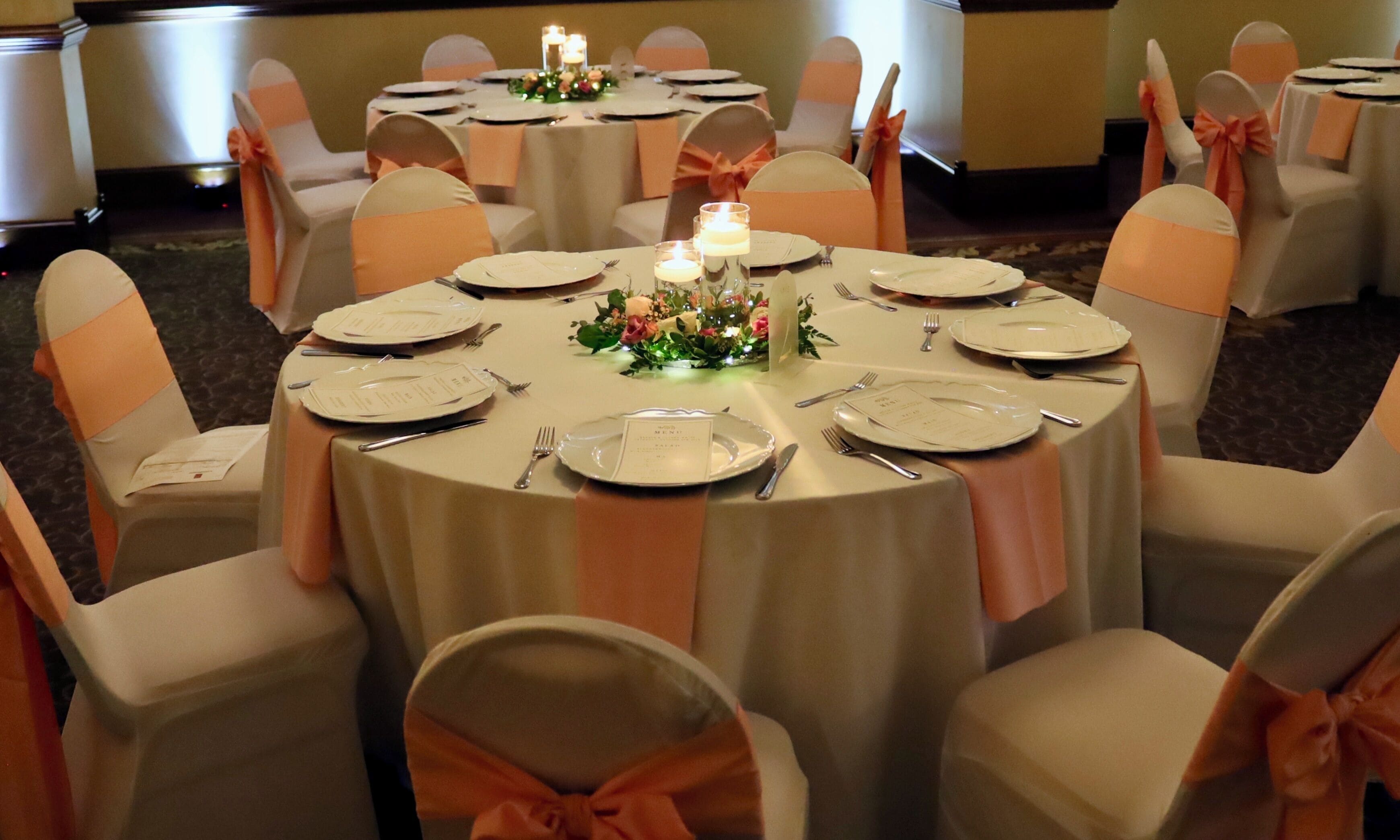 Une table ronde dressée pour un événement formel, avec des nappes blanches, des chaises assorties et des centres de table floraux.