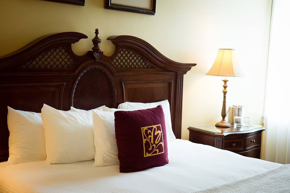 Una cama bien tendida con almohadas blancas y granate, un cabecero de madera y una mesita de noche con una lámpara y vasos.