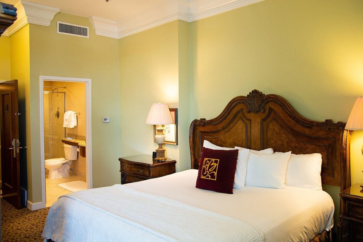 Una habitación de hotel con una cama grande, cabecera de madera, mesita de noche con lámpara, baño con ducha y puerta que da a otra habitación.