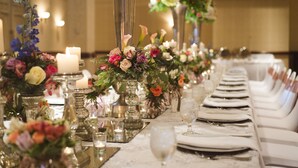 Una larga mesa de comedor con centros de mesa florales, velas y vajilla blanca.