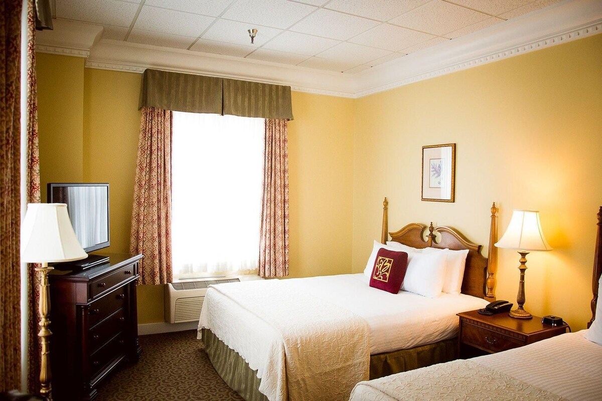 Habitación de hotel con cama, mesita de noche, lámpara, televisor, cómoda y ventana con cortinas.