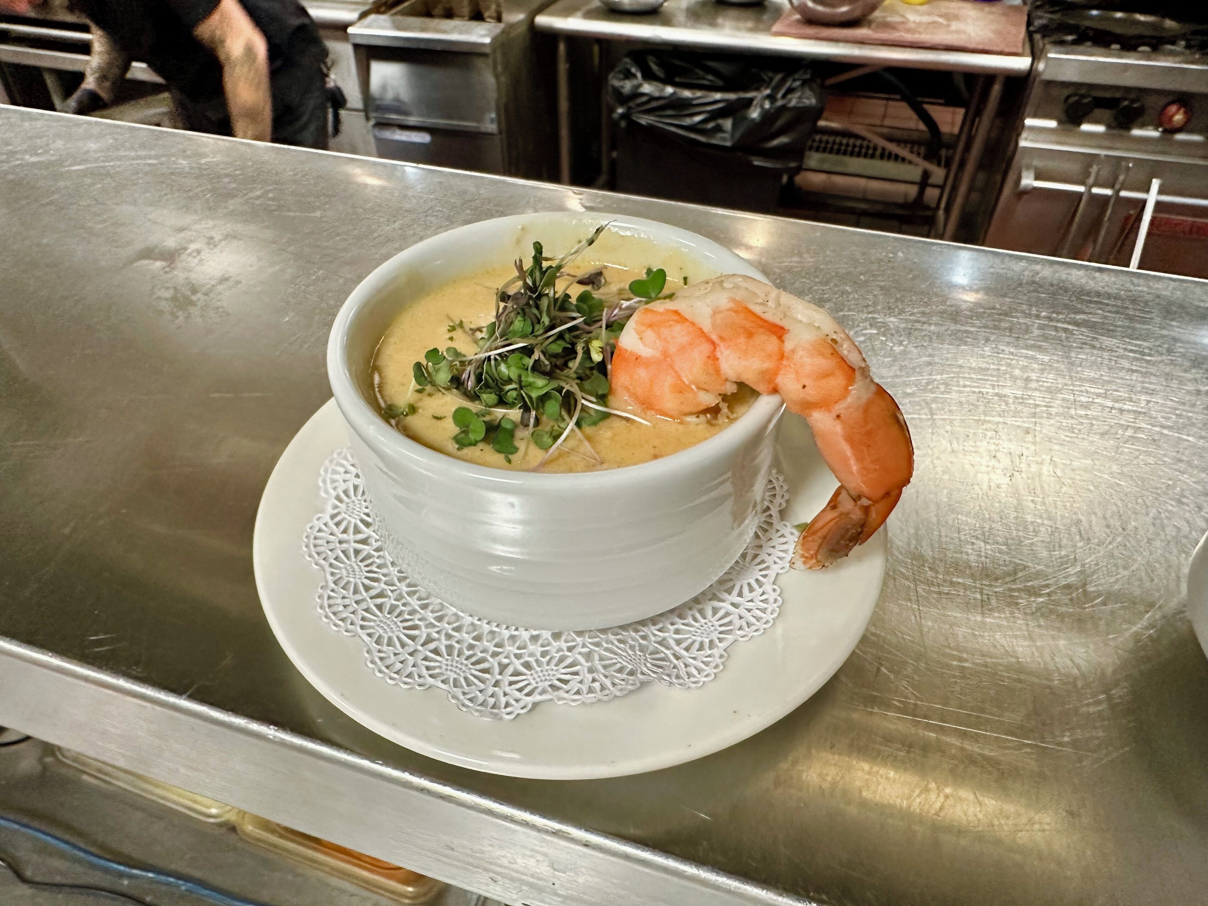 A bowl of soup with a shrimp garnish on a white plate with a lace doily, placed on a stainless steel table.