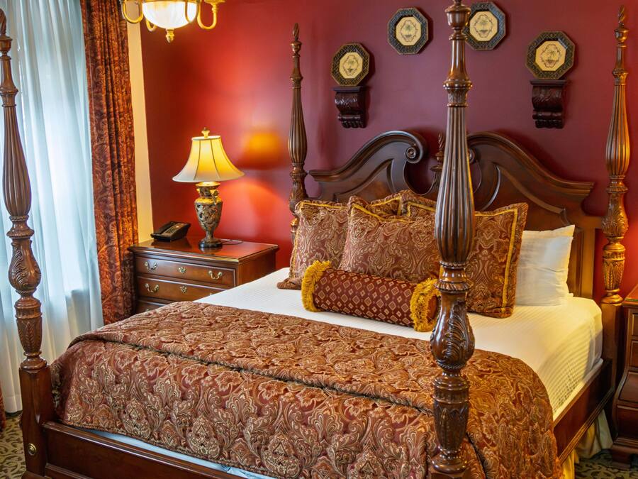 Una cama con dosel y un tablero de cabecera ornamentado, una mesita de noche con lámpara y un reloj en la pared.