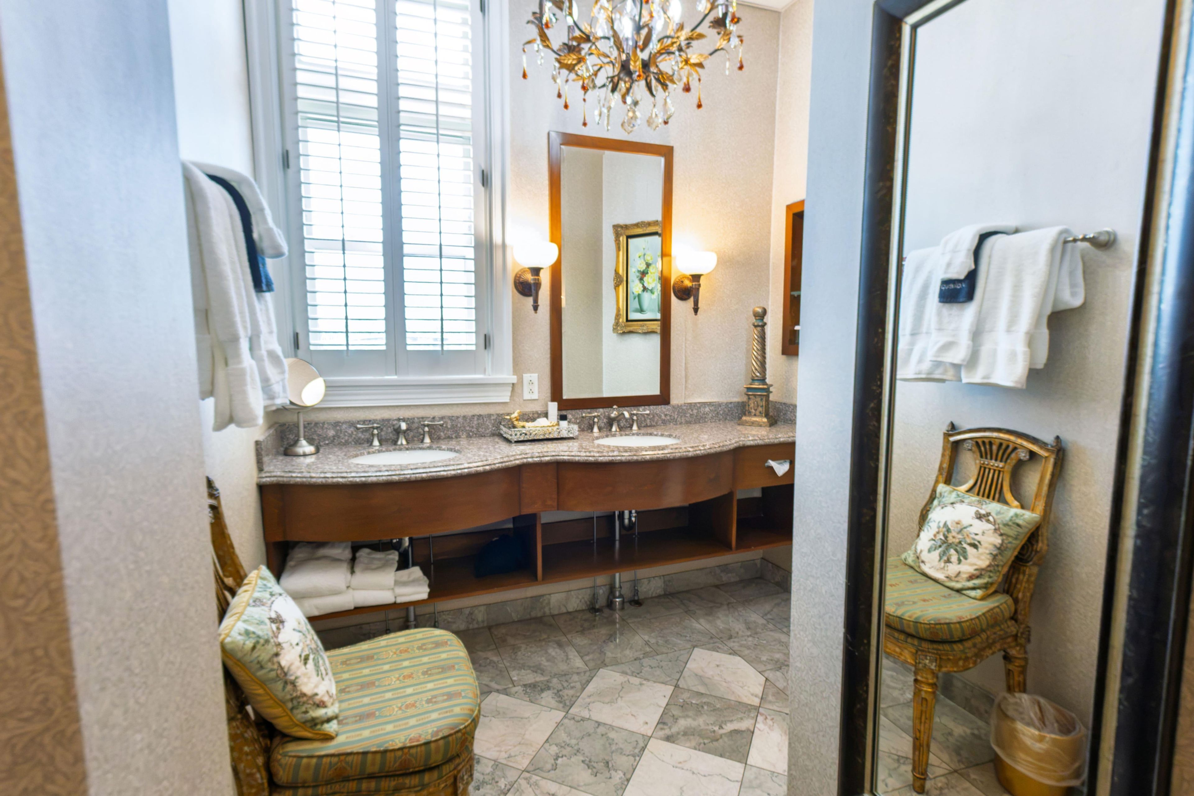 Un baño con tocador doble, espejo, candelabro, una silla y ventana con persianas.