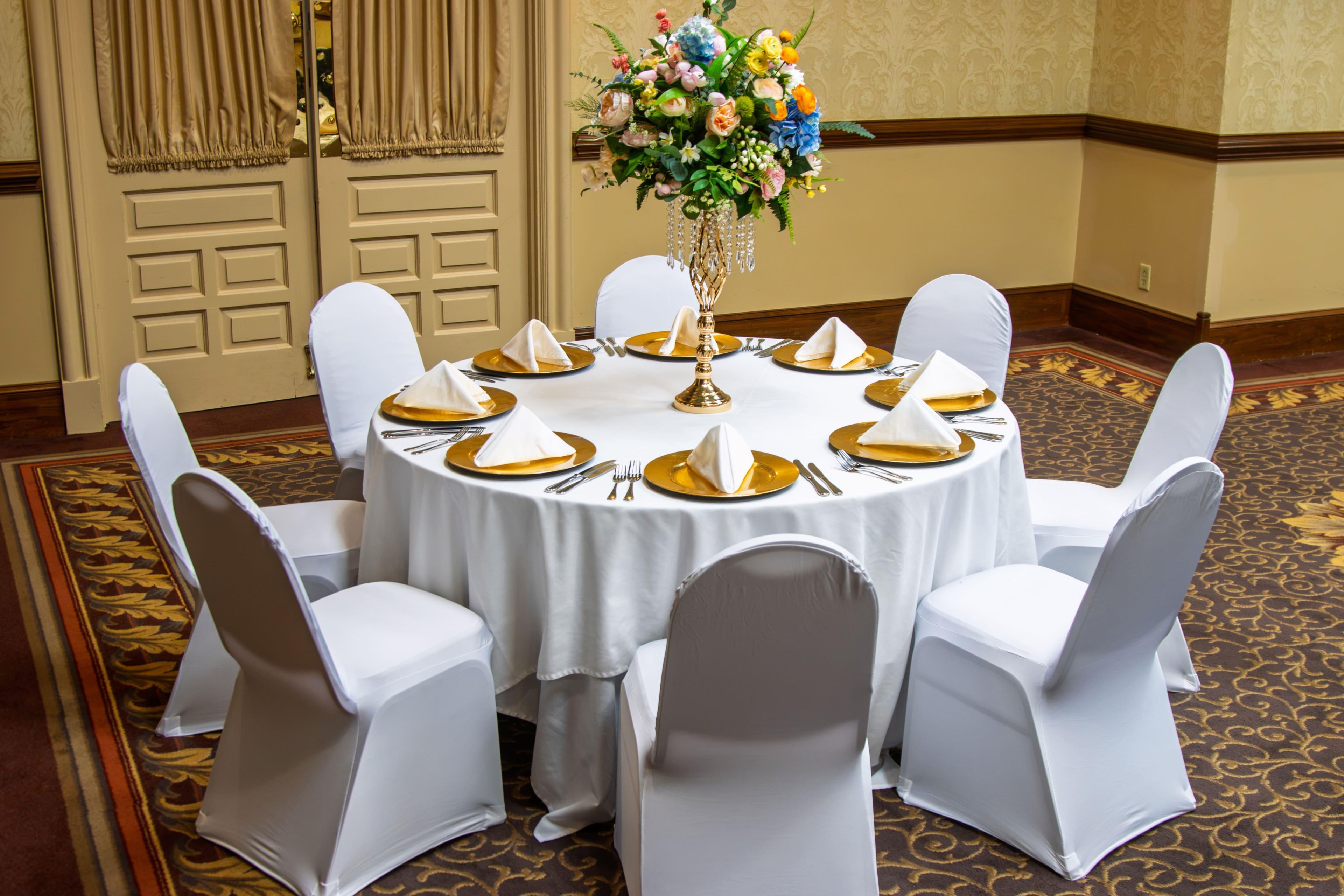 A round table set for six with white chairs, gold chargers, and a centerpiece of flowers.