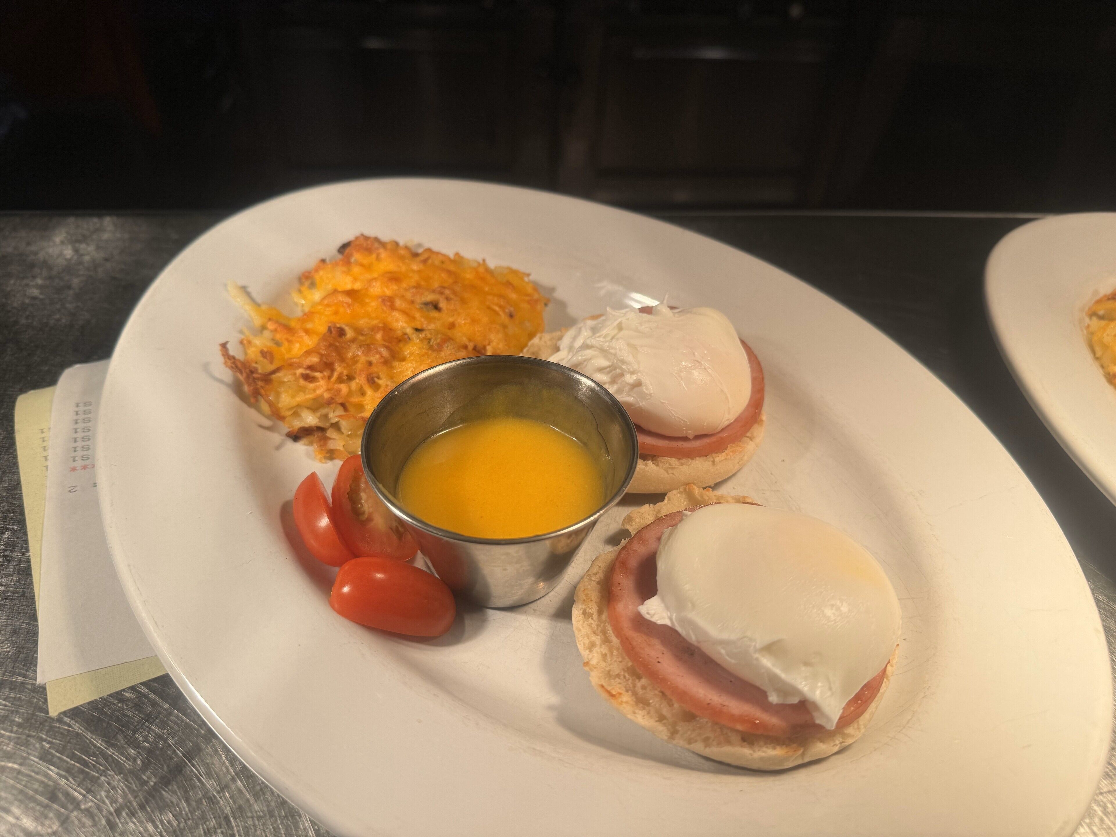 A plate with a ham and egg sandwich, a fried egg, hash browns, and a small metal cup of egg yolk.