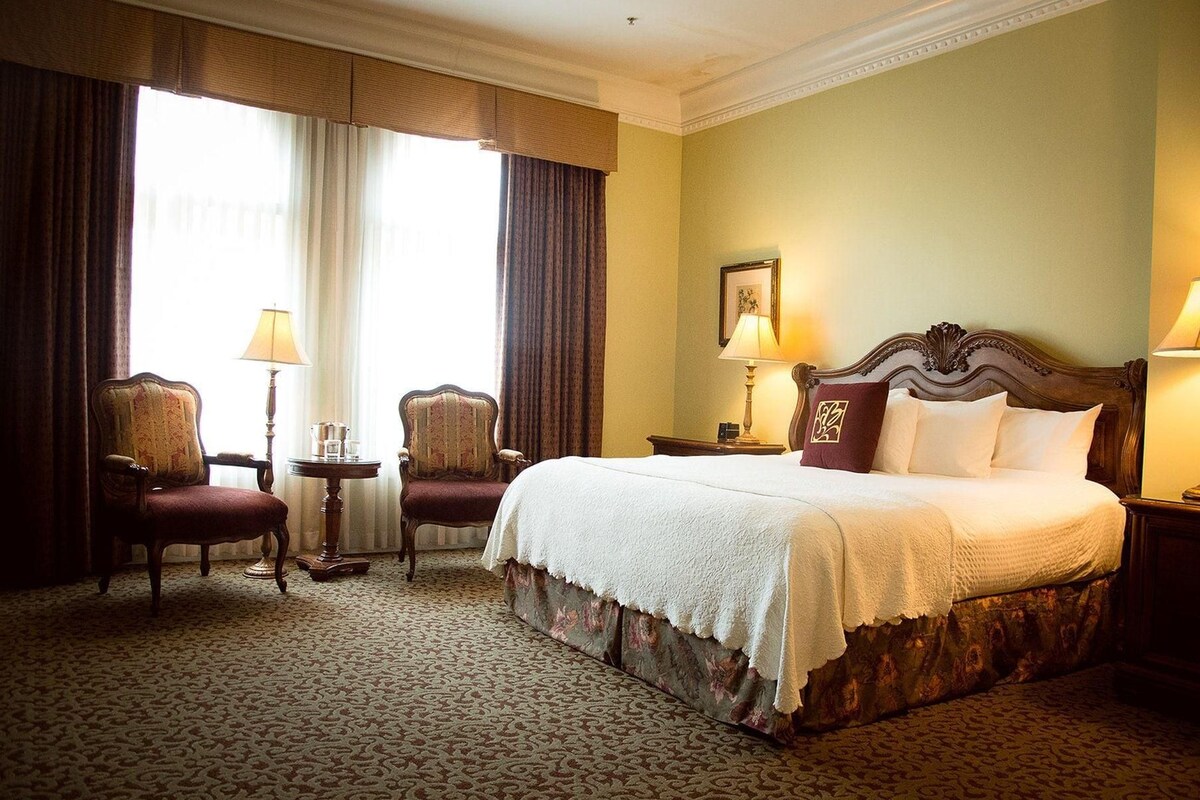Una habitación de hotel con una cama grande, dos sillones, una mesita y una mesita de noche con lámparas.