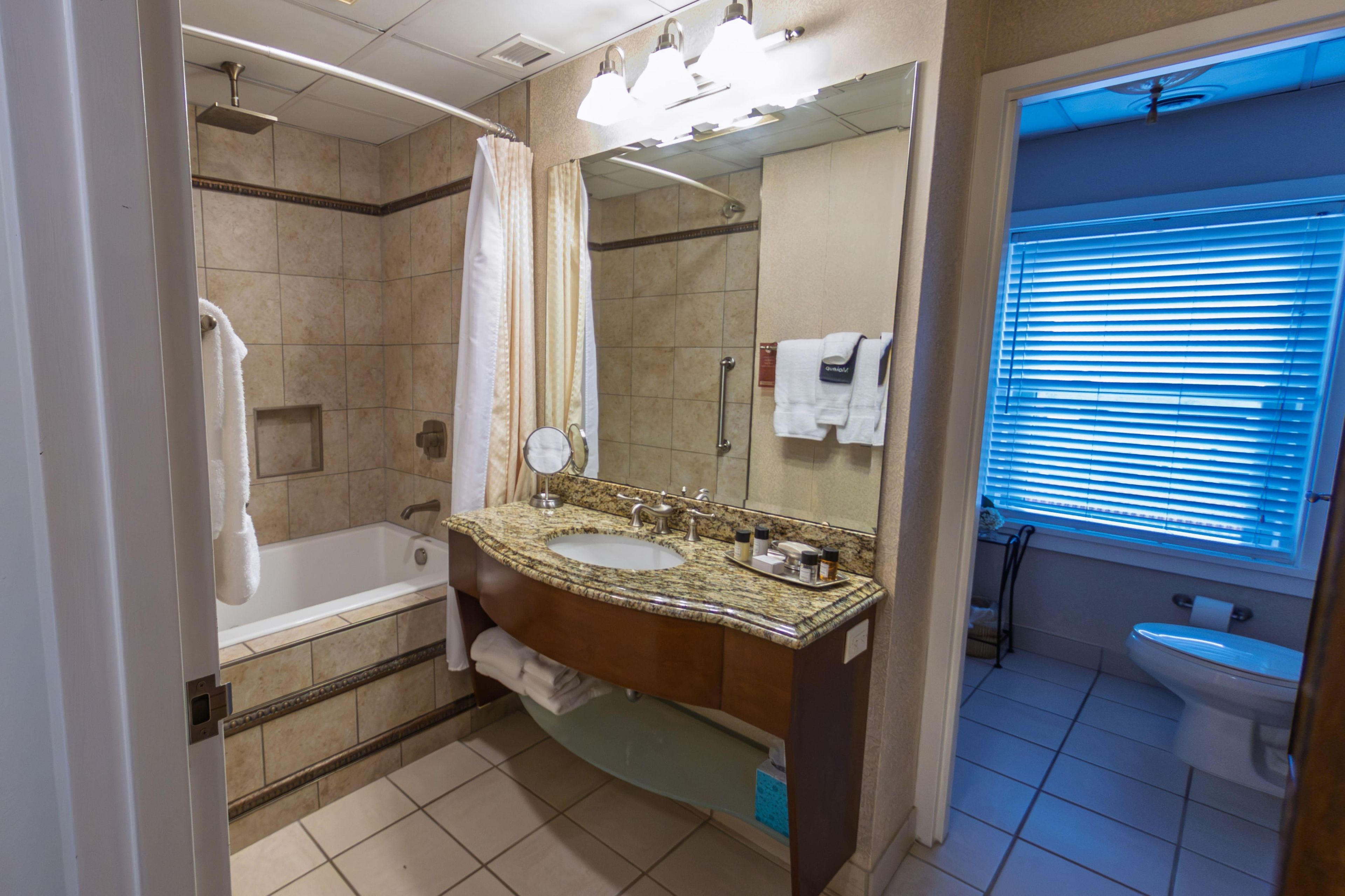Un baño con bañera, lavamanos con tocador, espejo y ventana con persianas.