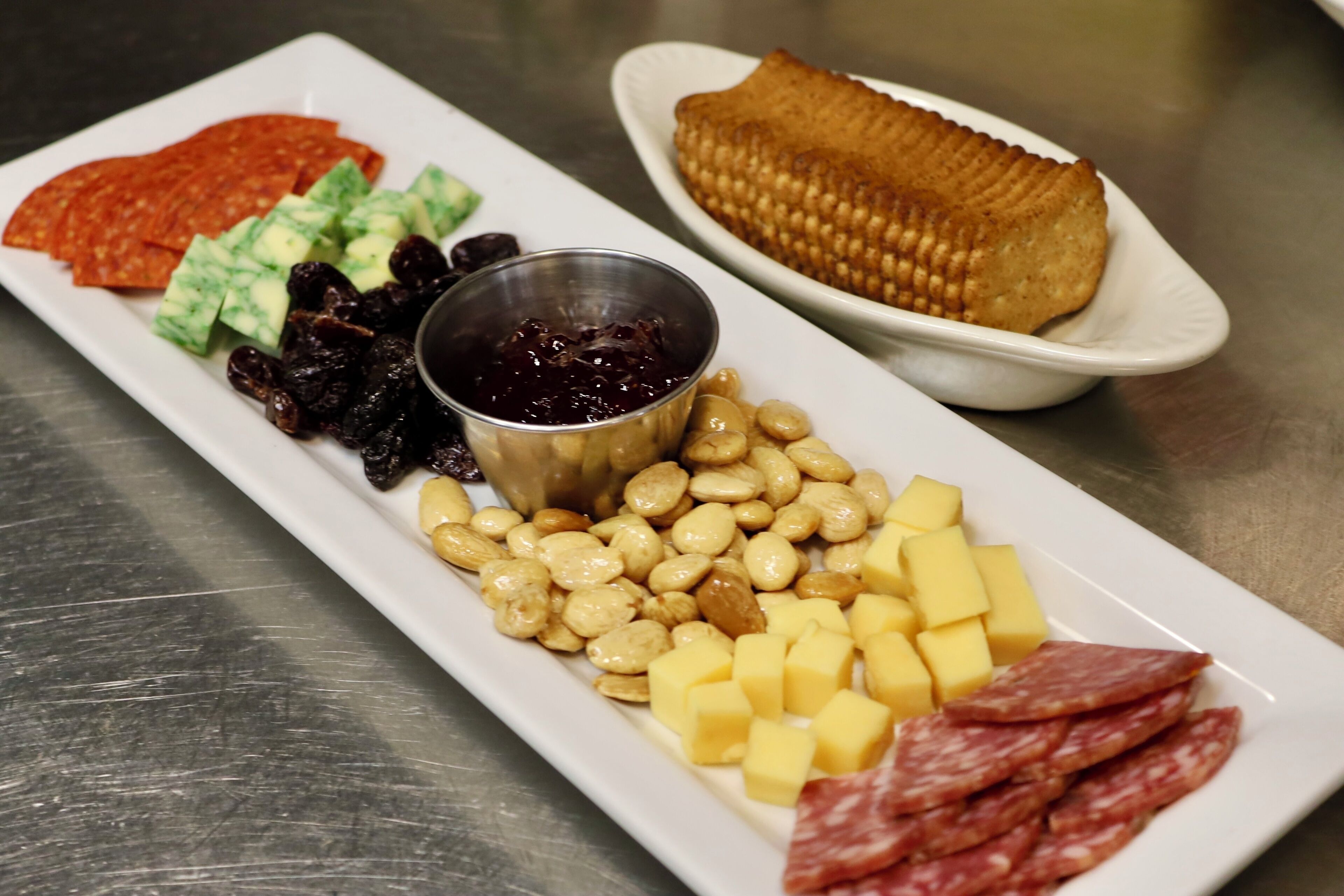 A white rectangular plate with various food items including cheese, nuts, dried fruits, and a small bowl of jam.