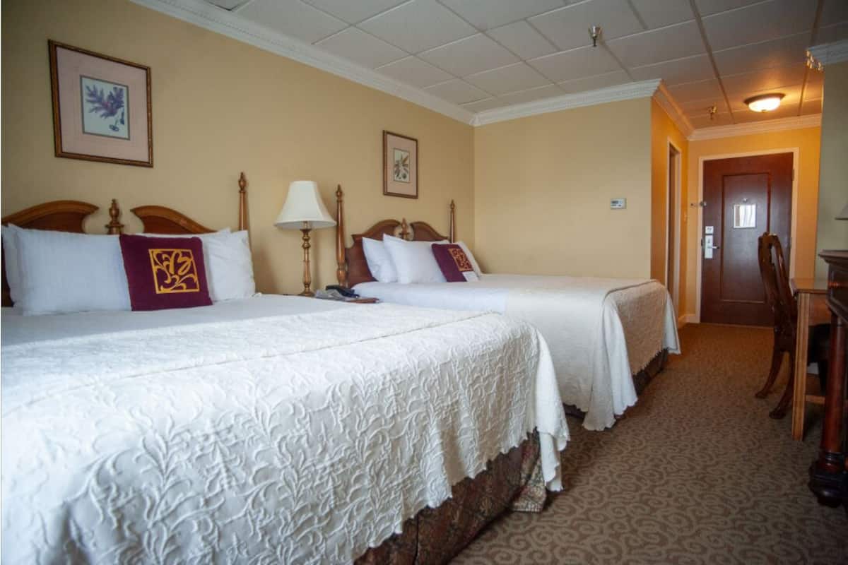 Habitación de hotel con dos camas, cada una con cubrecama blanco y almohada decorativa. Se observa un cabecero de madera, una lámpara de noche y un cuadro enmarcado en la pared. El suelo está alfombrado y hay una puerta que conduce a otra zona.