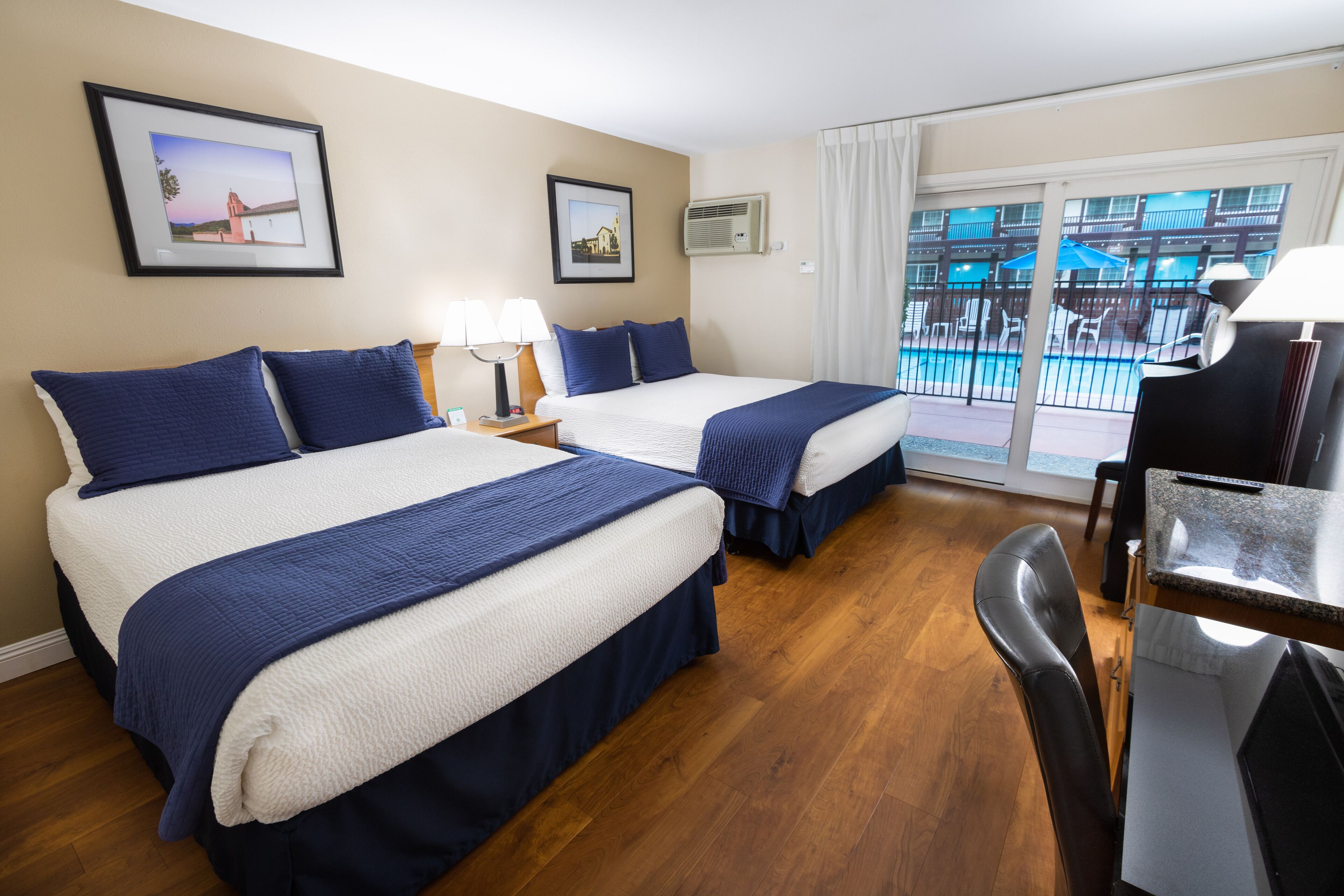 A hotel room with two beds, a desk, a chair, and a balcony with a view of a building and a pool.