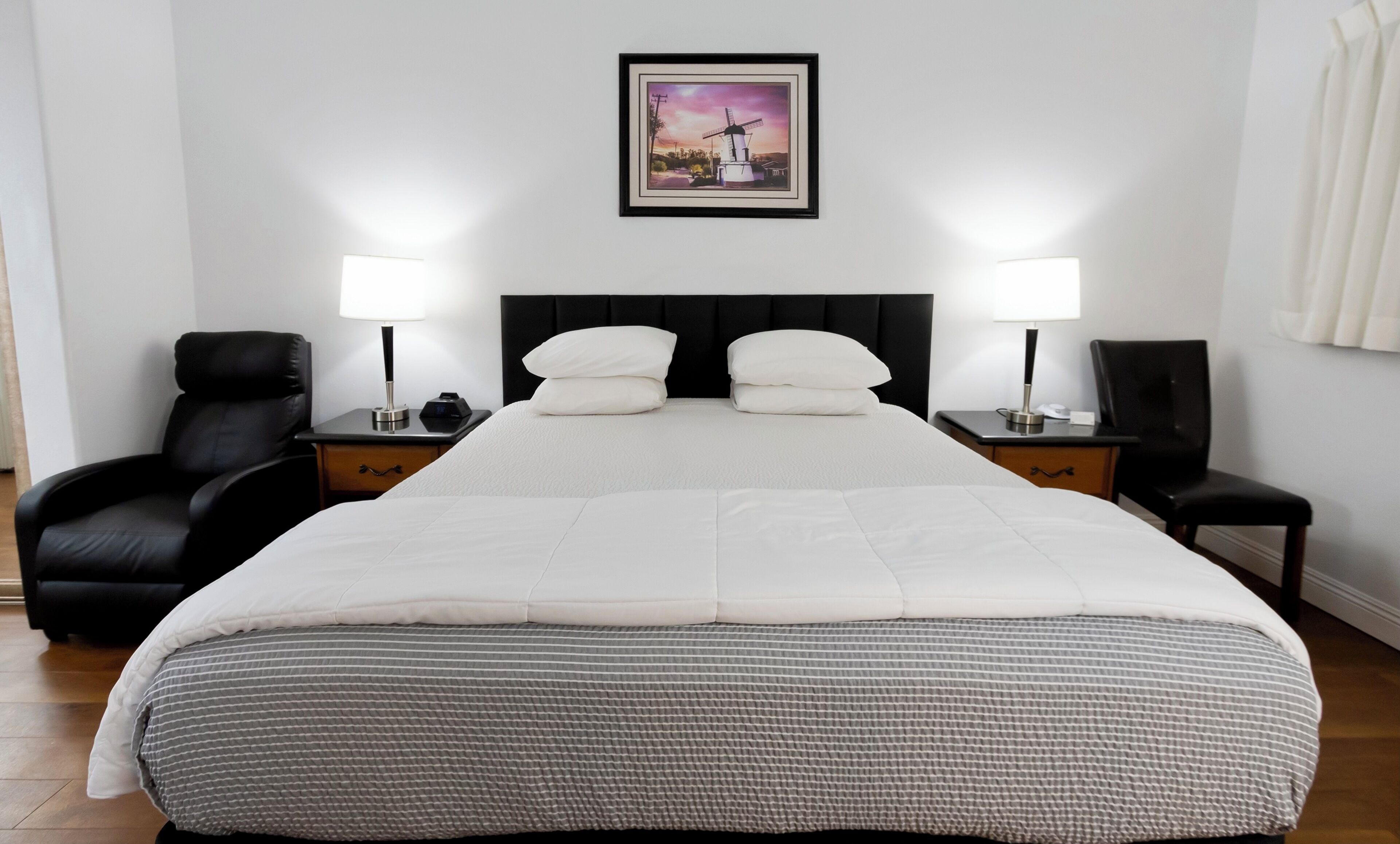 A hotel room with a large bed, two bedside tables, a black leather chair, and a framed picture on the wall.