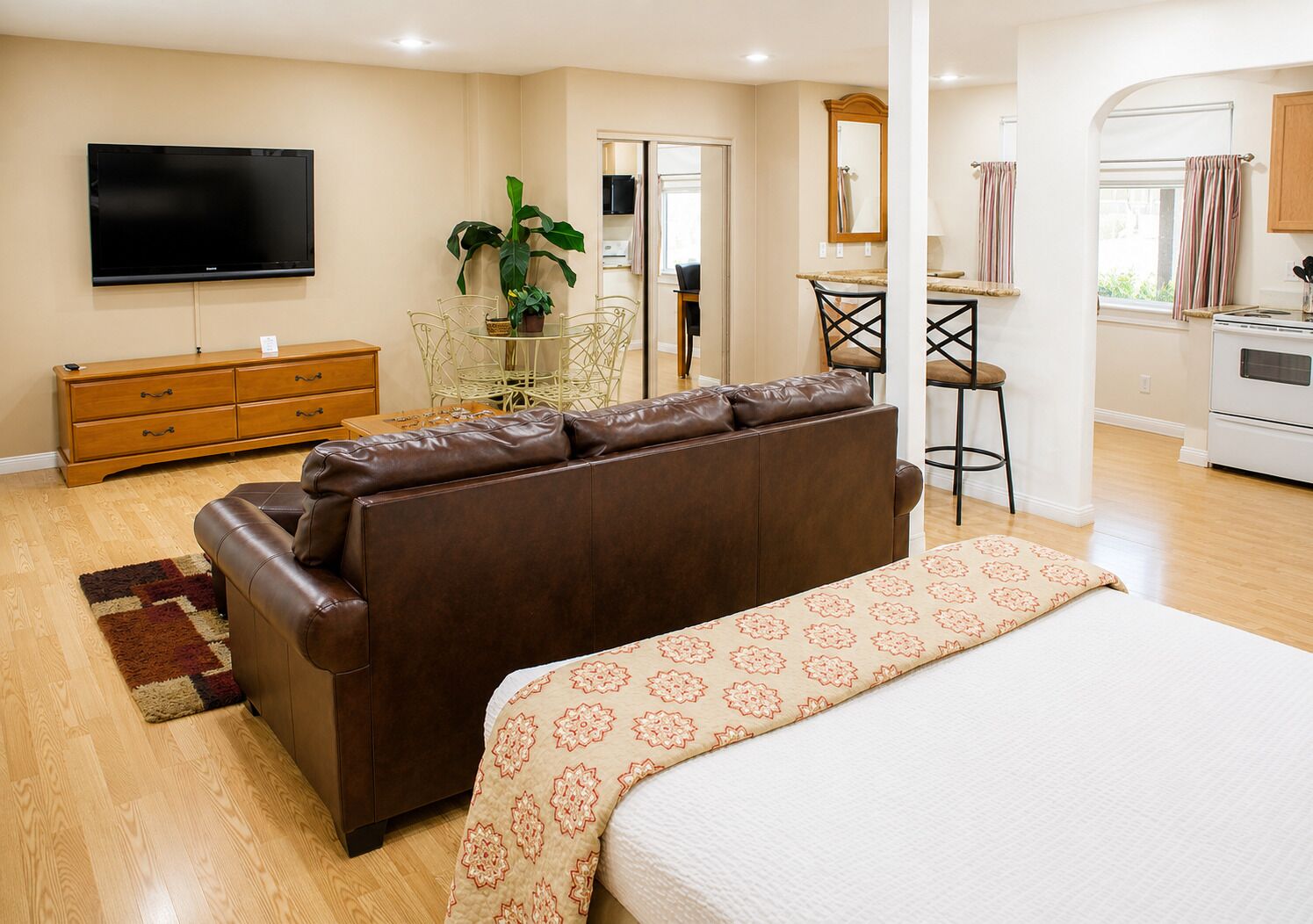 Un dormitorio con una cama grande, un sofá de cuero marrón, un televisor, un aparador de madera, una mesa de comedor con sillas y una zona de cocina.