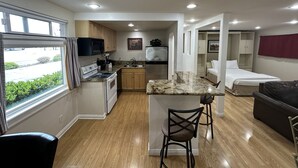 A compact kitchen with a stove, microwave, and sink, a small dining area with a bench, and a bedroom with a bed and a wardrobe.