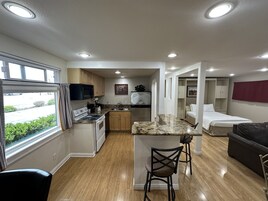 Cocina compacta con estufa, microondas y lavamanos, un rincón comedor con un banco, y un dormitorio con cama y armario.