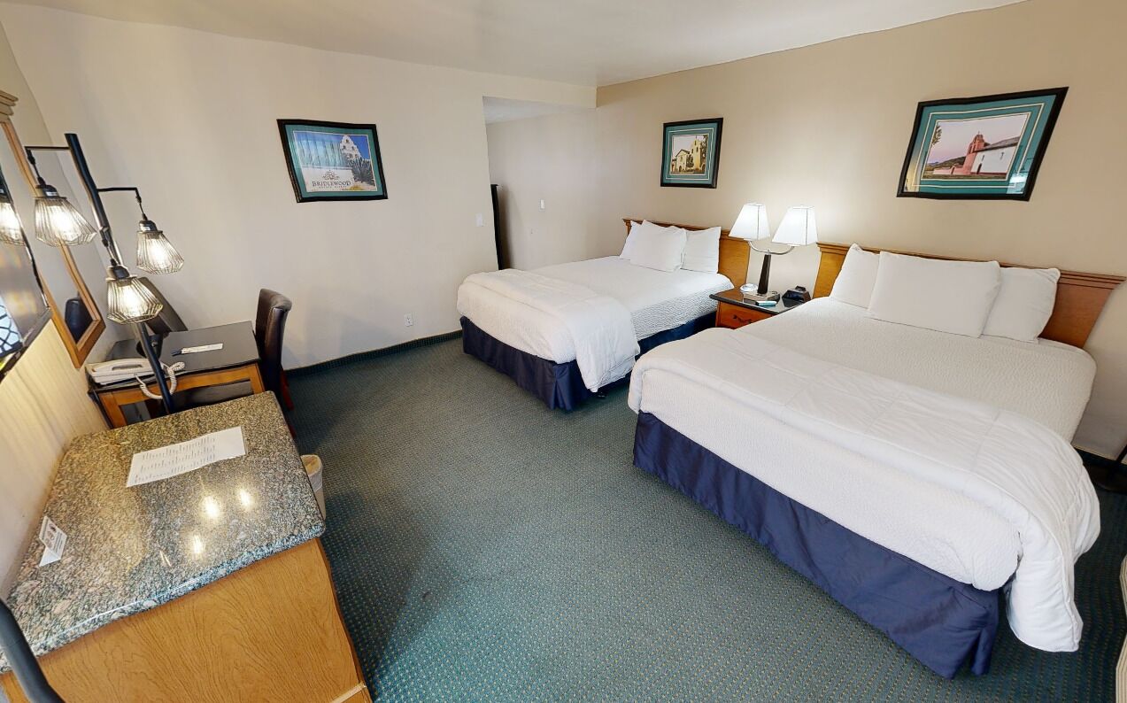 A hotel room with two beds, a desk, and a chair. There are wall-mounted lamps, a small table, and framed pictures on the walls.