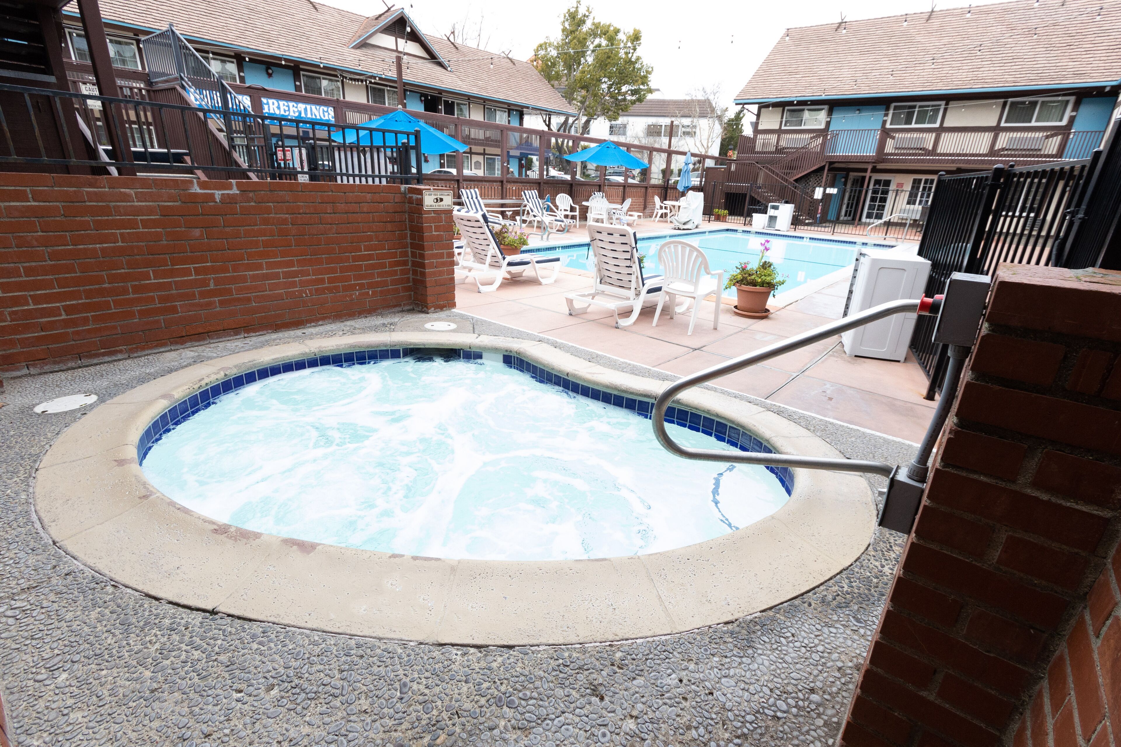 A hot tub surrounded by lounge chairs and a pool, with a brick wall and a building in the background.