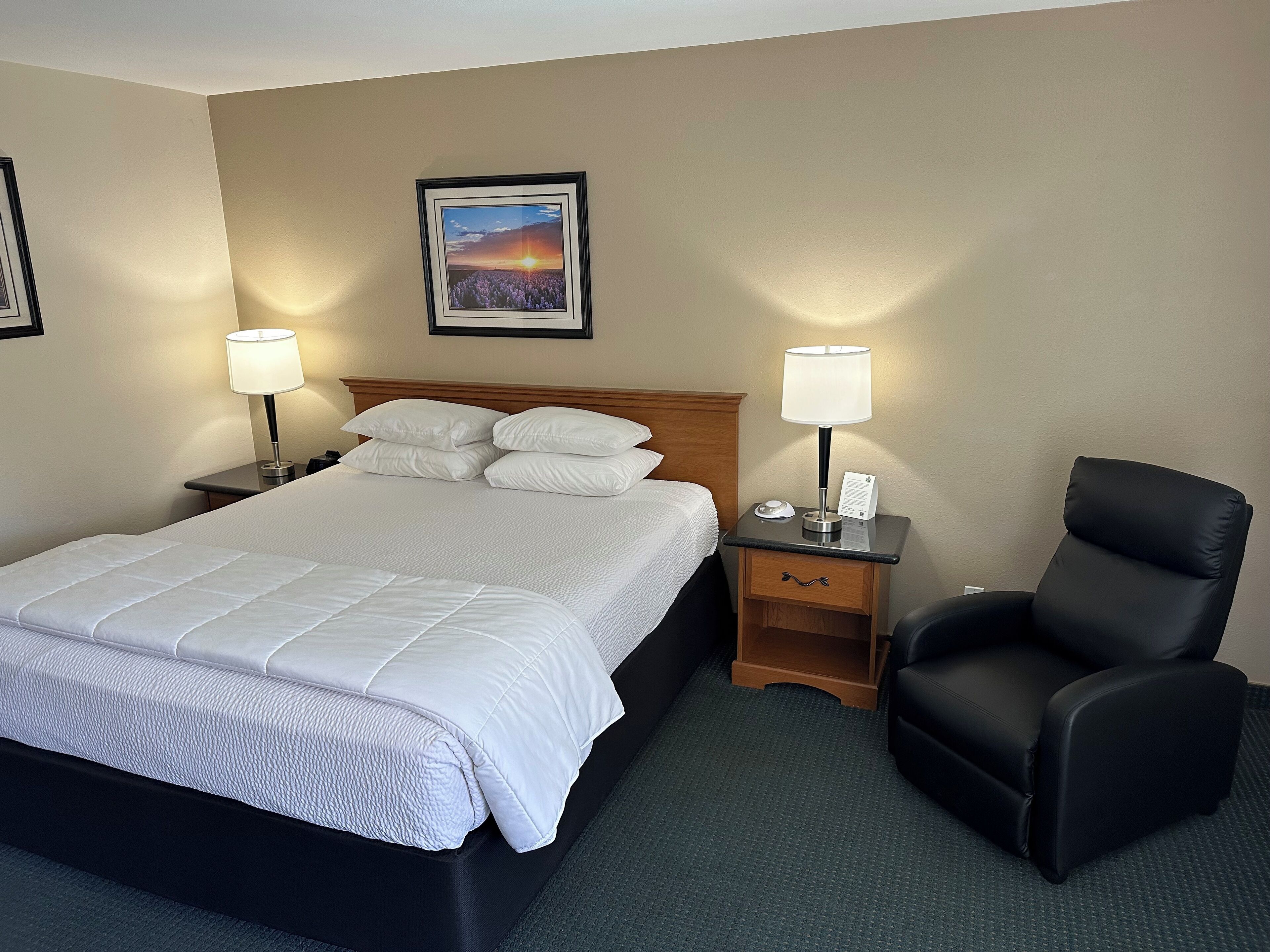 Una habitación de hotel con una cama, dos mesitas de noche con lámparas, una silla y un cuadro en la pared.