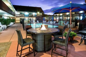 An outdoor pool area with a fire pit table, wicker chairs, and umbrellas. - King Frederik Inn (Solvang)