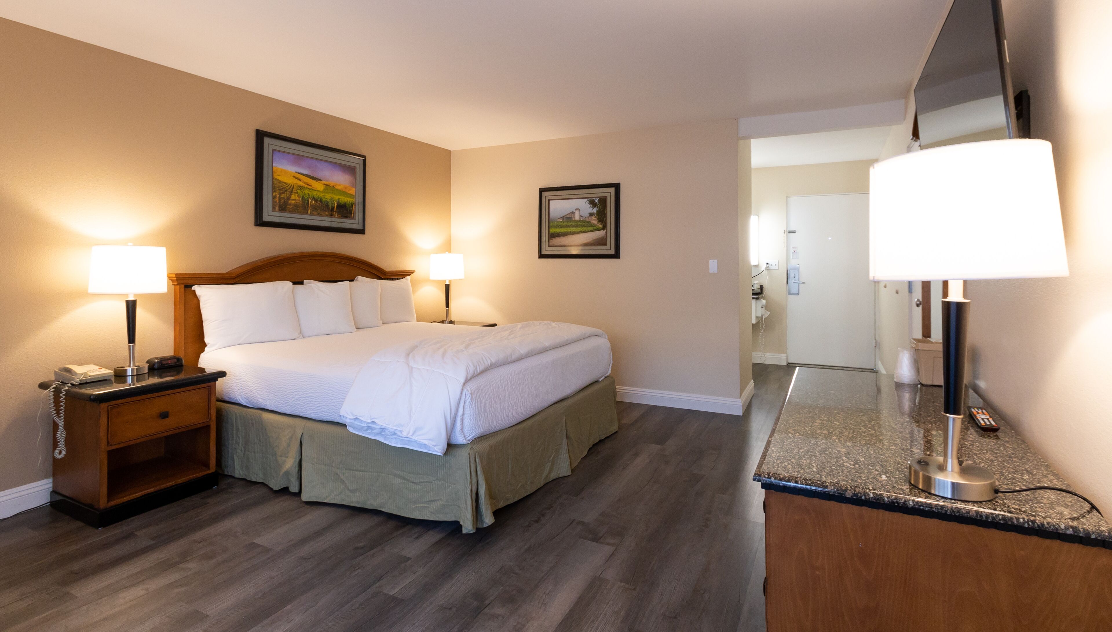 Een hotelkamer met een groot bed, twee bedlampjes, een nachtkastje, een dressoir, een spiegel en twee ingelijste foto’s aan de muur.