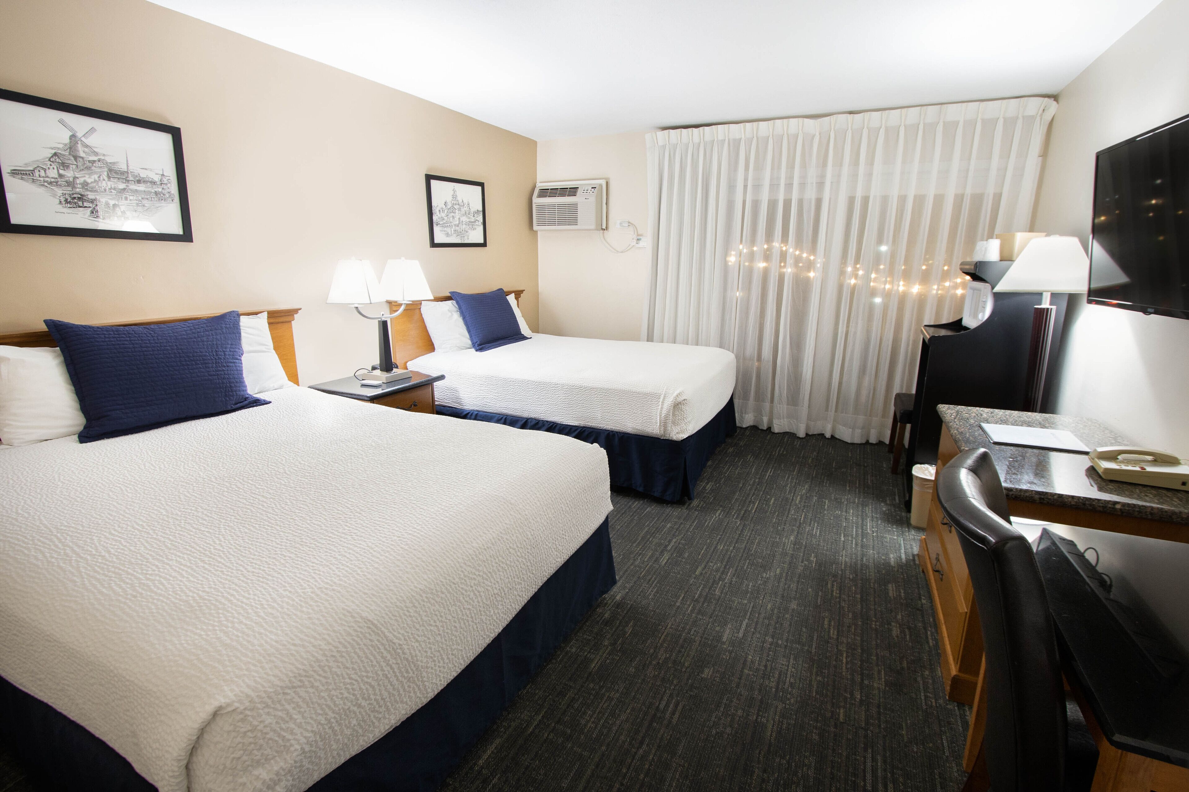 Quarto de hotel com duas camas, uma escrivaninha, uma cadeira, televisão e janela com cortinas.
