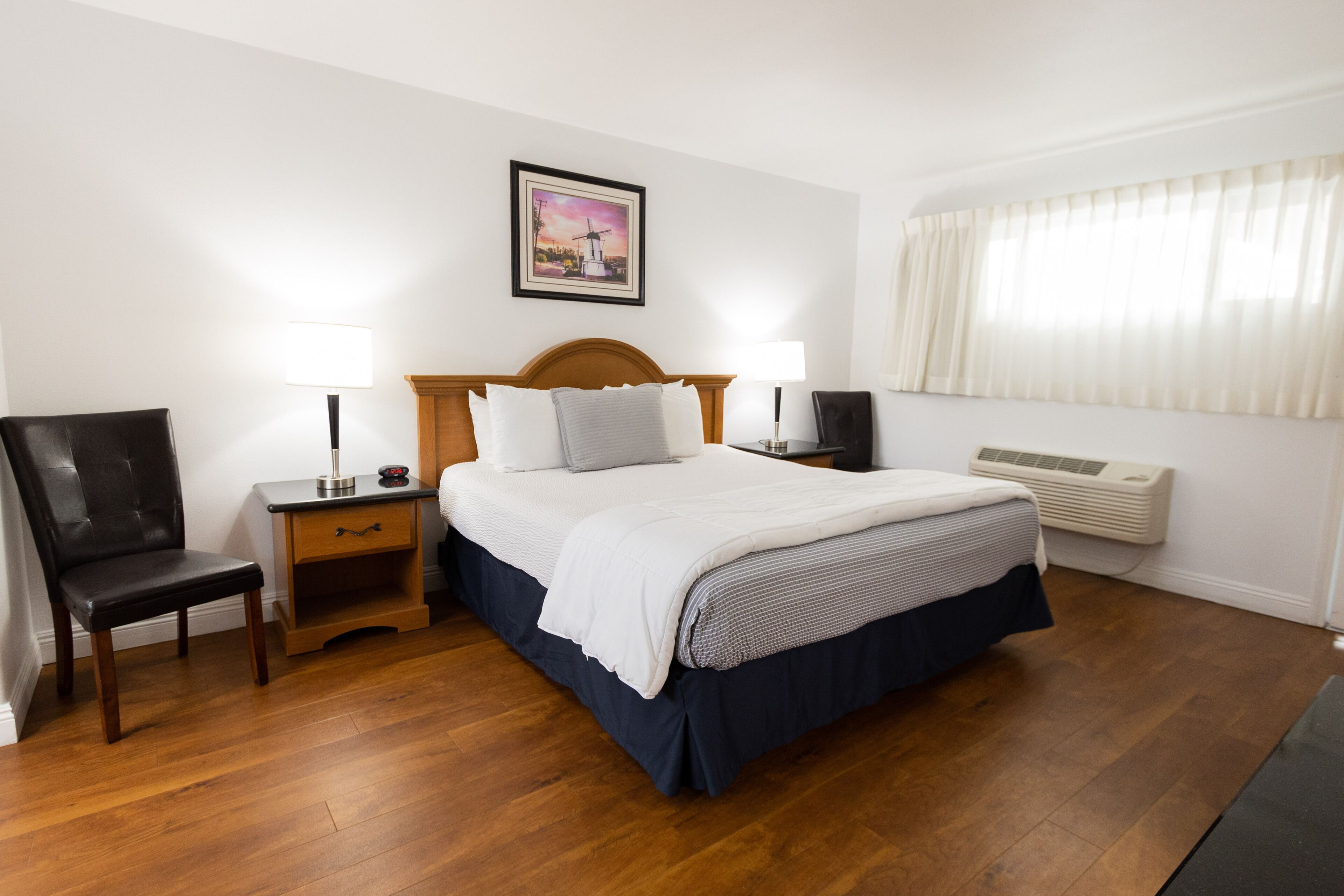 Une chambre d’hôtel avec un lit en bois, deux tables de chevet avec des lampes, une chaise, un tableau au mur et une fenêtre avec des rideaux.