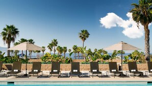 Un espace aménagé au bord de la piscine, avec des chaises longues et des parasols, des palmiers et une vue sur l’océan.