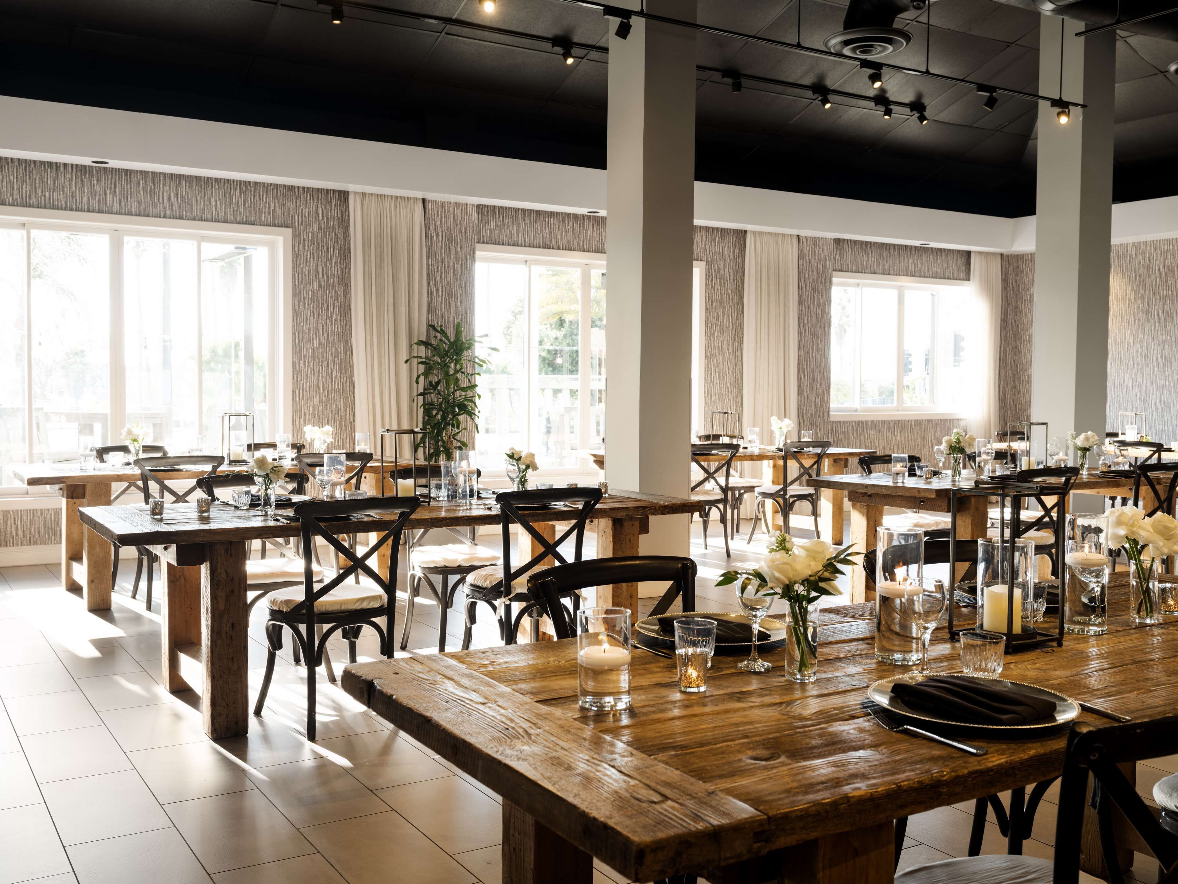 A spacious dining area with multiple wooden tables and chairs, large windows, and a modern ceiling design.