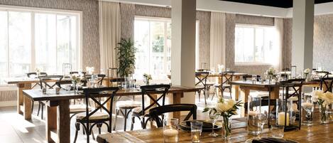 A spacious dining area with multiple wooden tables and chairs, large windows, and a modern ceiling design.