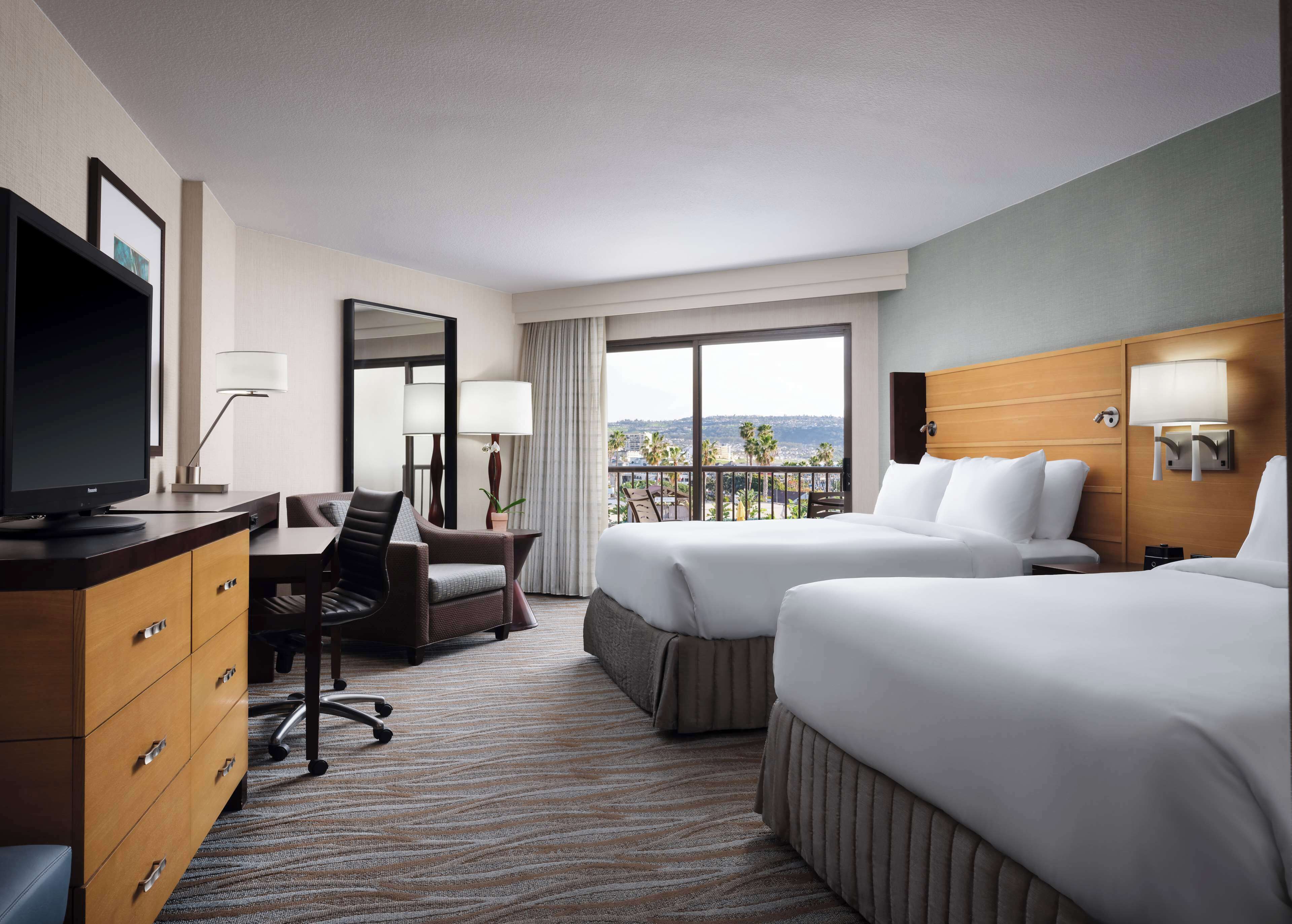 A hotel room with a desk, two beds, a TV, and a balcony with a city view.